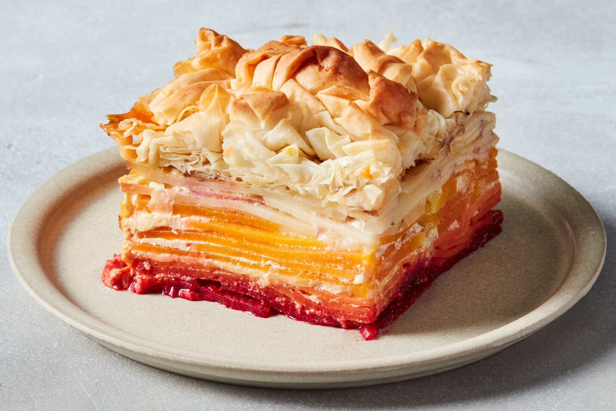 A side image of a layered gratin made of red beets, orange squash and white potatoes. Crinkled, burnished phyllo sheets sit on top.