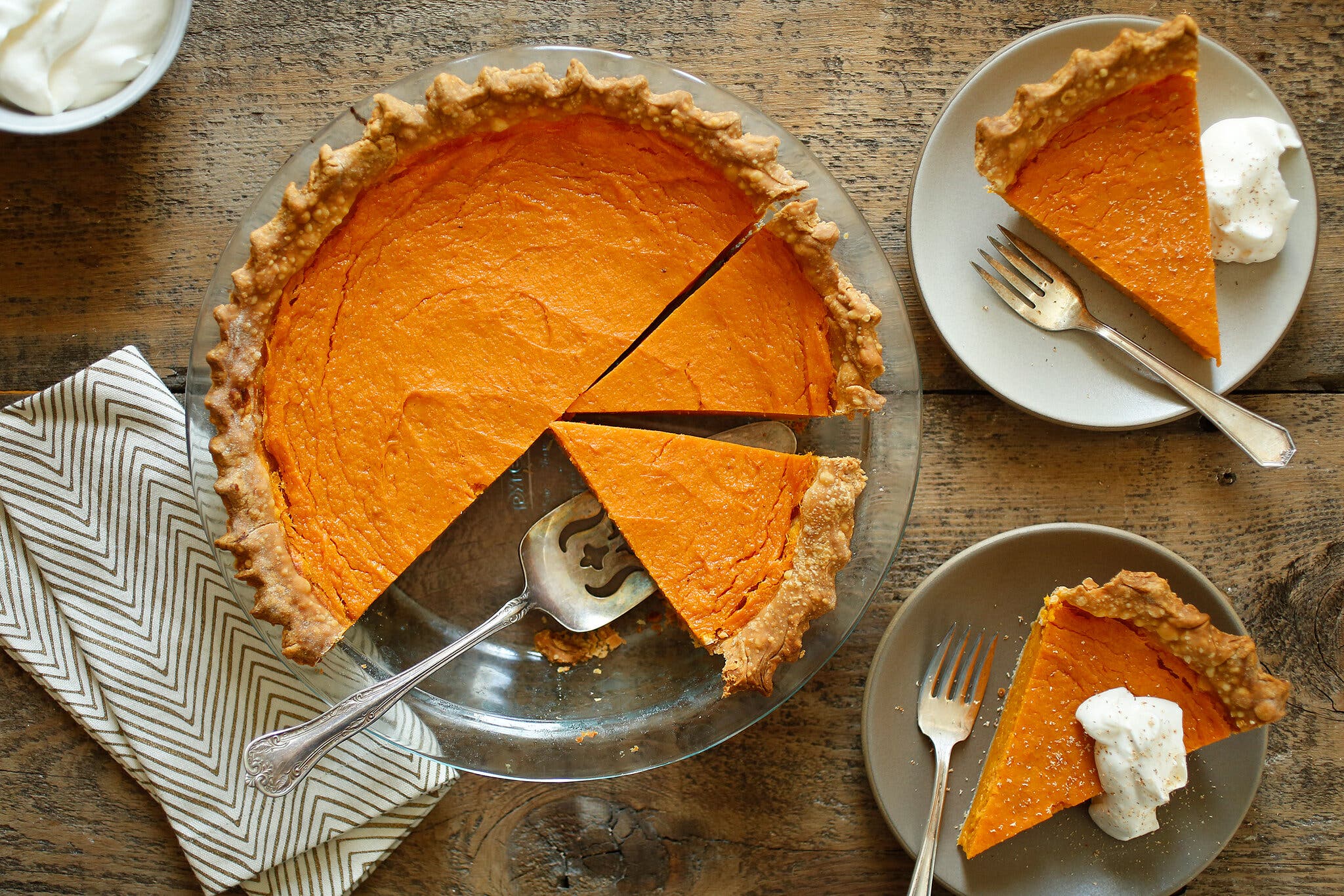 A bright-orange sweet potato pie in a glass pie dish sits on a wooden countertop. Two slices are off to the right on white plates. To the left of the dish is a folded fabric napkin.