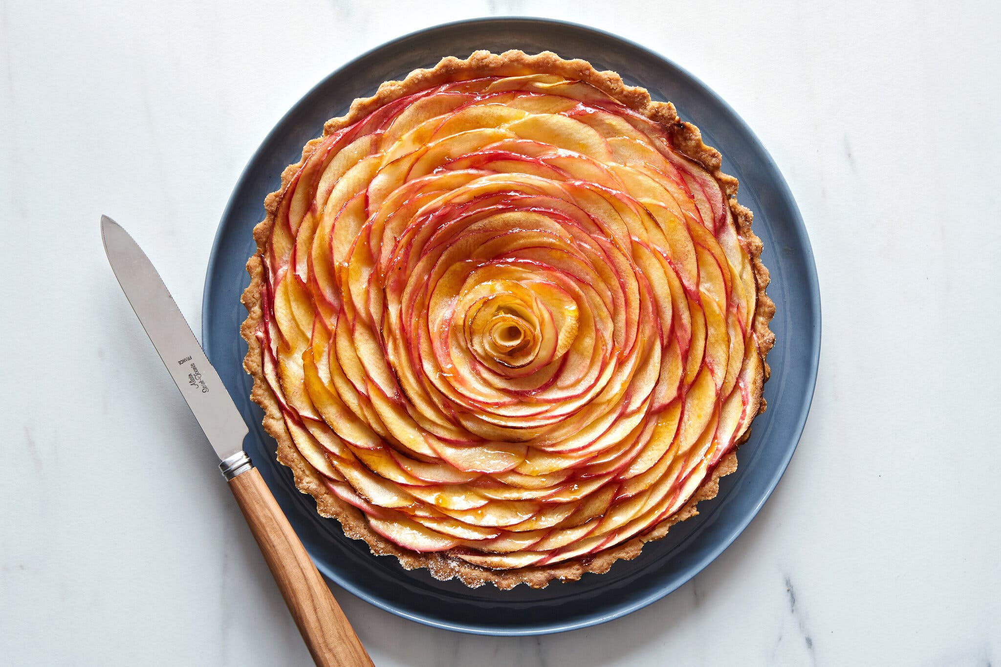 An apple tart, with thin apple slices arranged in the crust to look like a rose, sits on a blue plate against a marble background. A knife with a wooden handle is off to the left.