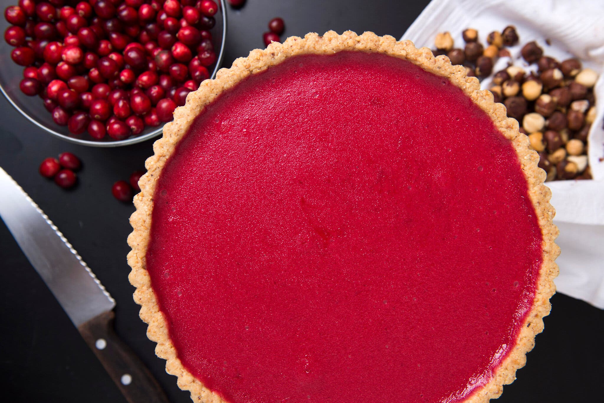 A bright-red cranberry curd tart, edged with a hazelnut crust, sits in the middle of the frame. In the background are a bowl with cranberries, a napkin holding hazelnuts and a serrated knife.