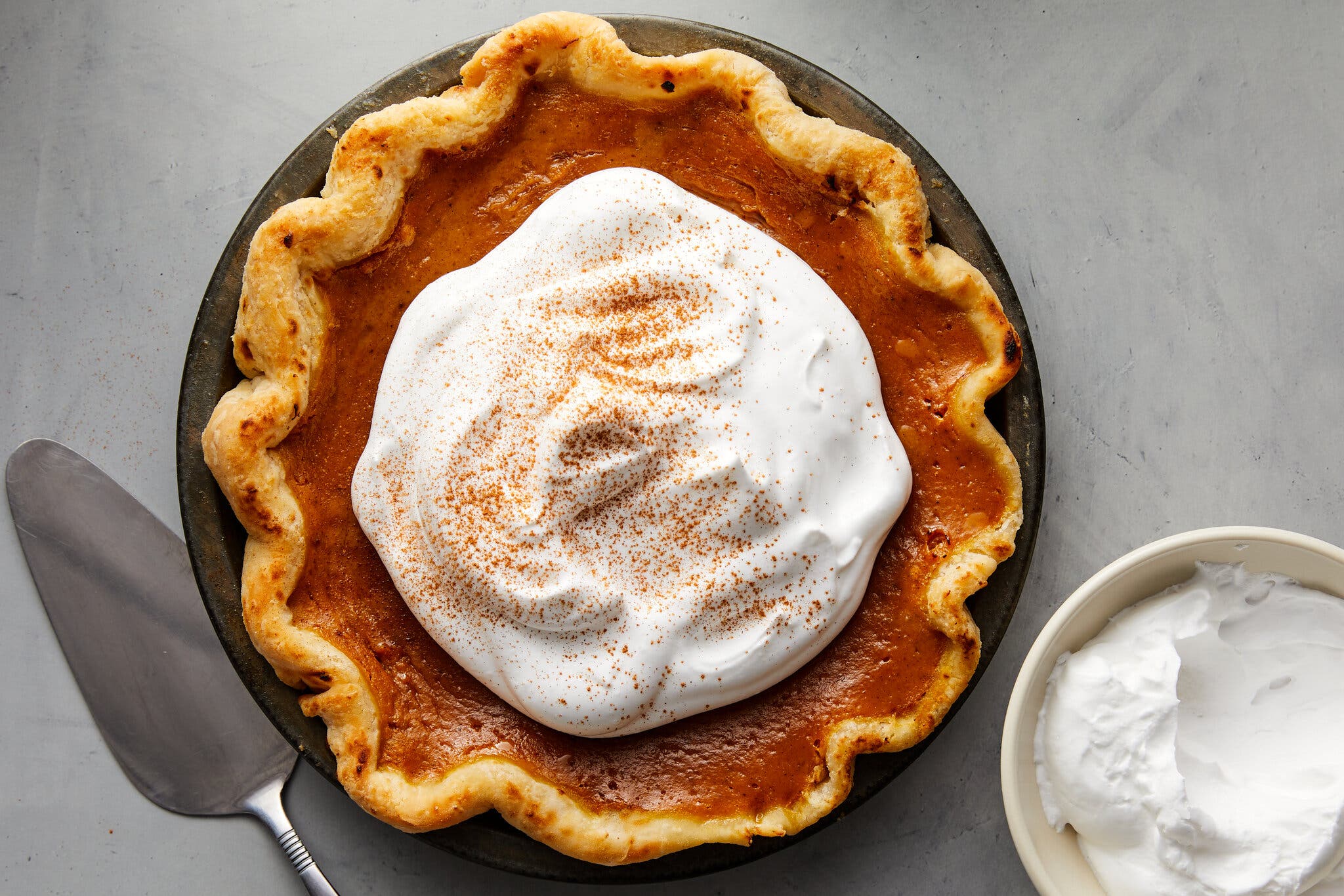 A pumpkin pie topped with whipped cream is shot from overhead and sits against a light gray background. To the left of the pie is a cake knife and to the right is a small bowl full of whipped cream.