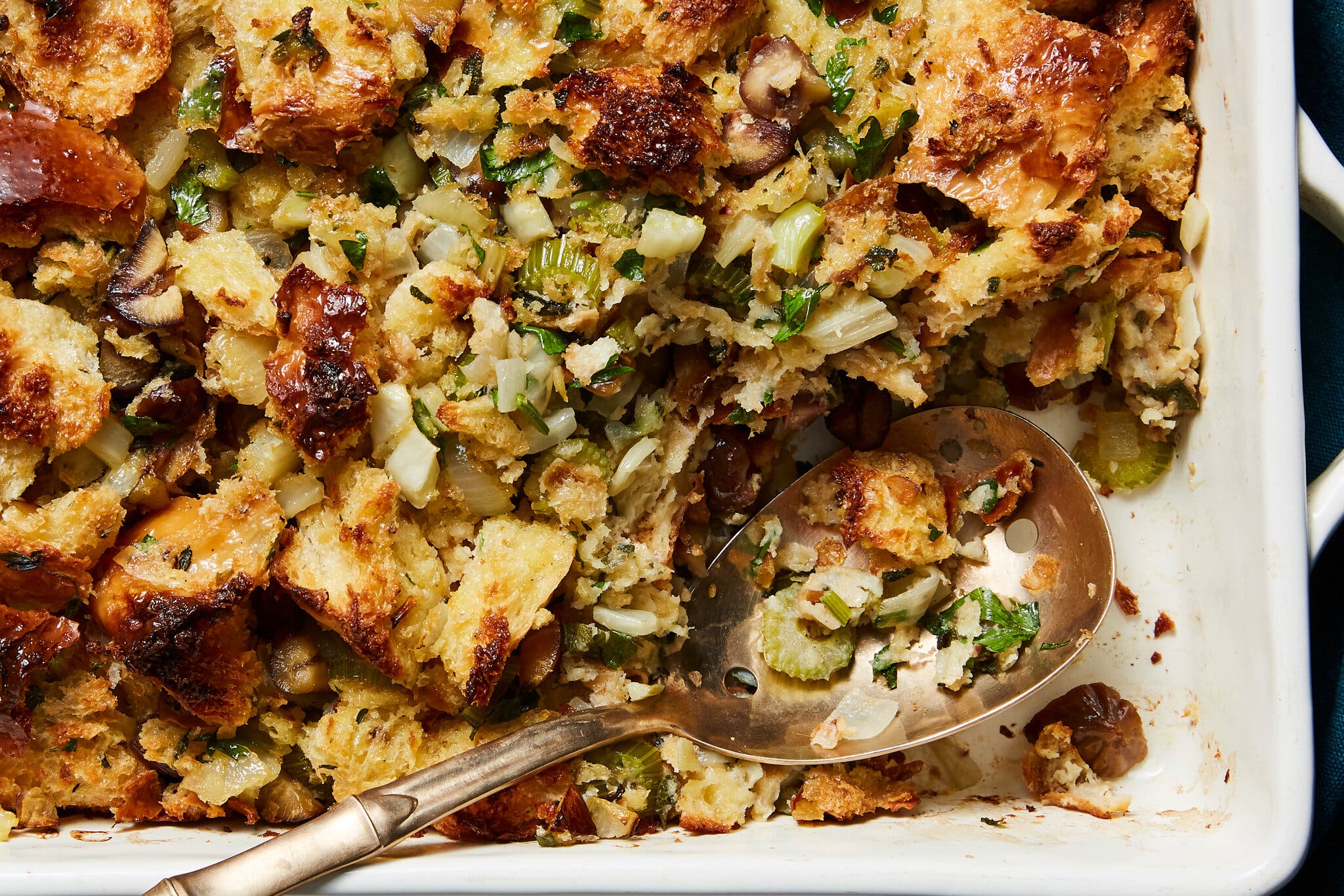 A large casserole dish holds a burnished chestnut stuffing. A portion has been taken out where a serving spoon now sits. To the right is a crumpled blue linen napkin.