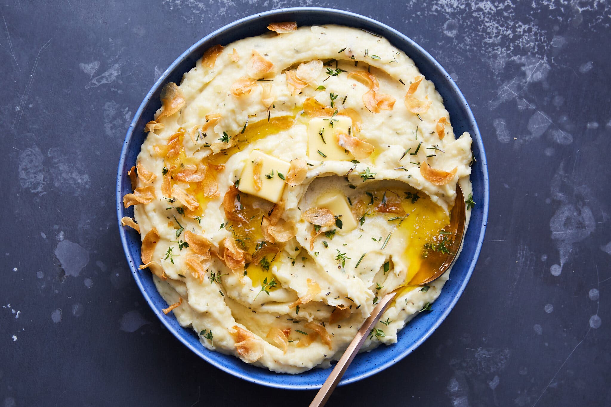 A blue bowl holding mashed potatoes topped with butter and crispy shallots sits against a dark blue background.