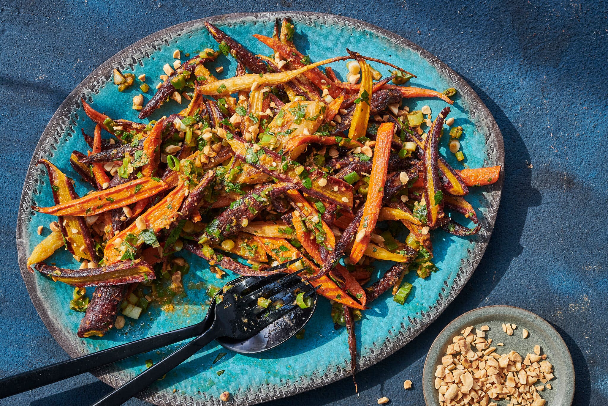 A blue plate with a gray rim is topped with sliced, multicolor carrots and yaji spice. To the bottom right is a small round plate holding crushed peanuts.