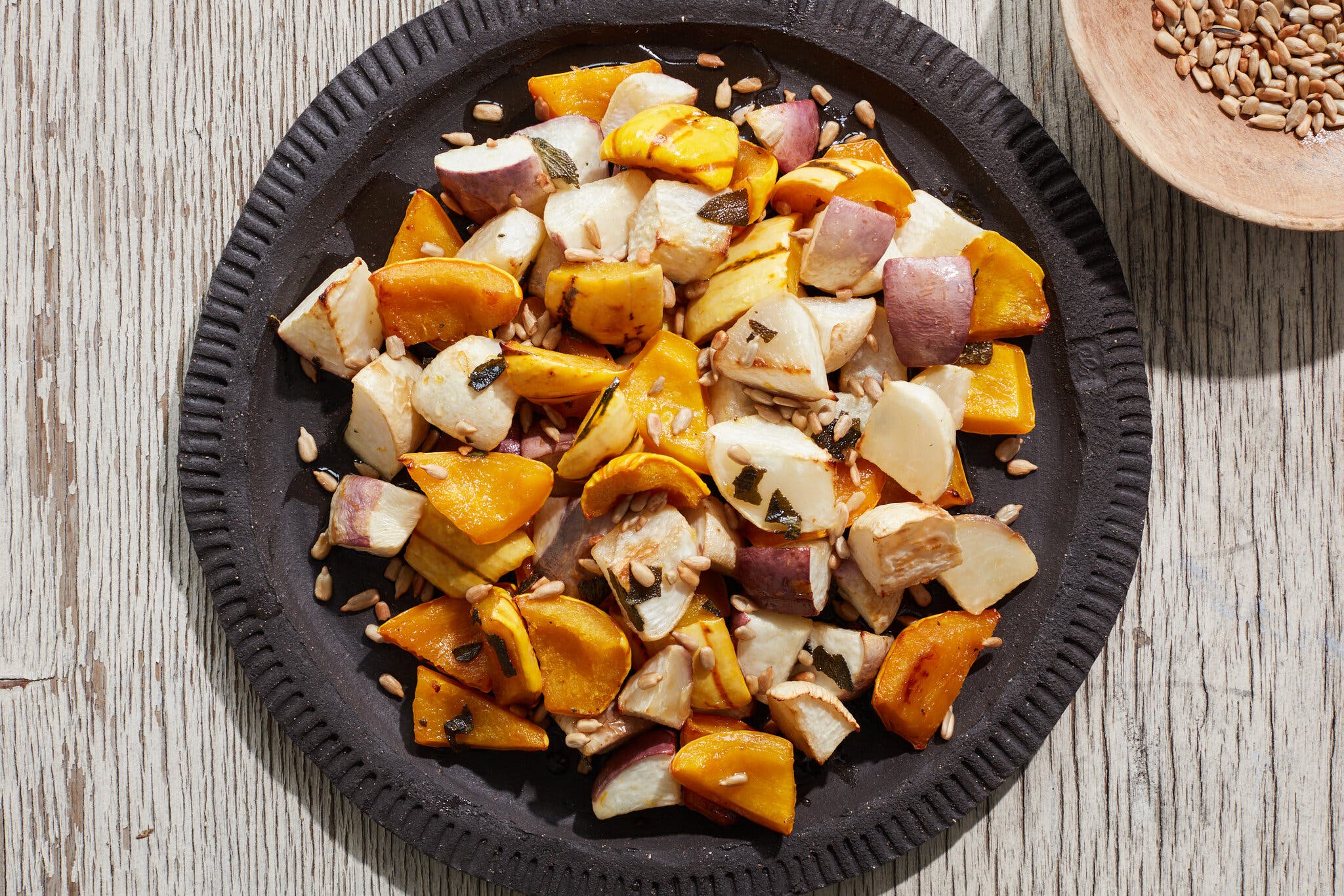 An overhead shot of assorted turnips and winter squash on a black plate, topped with pine nuts.