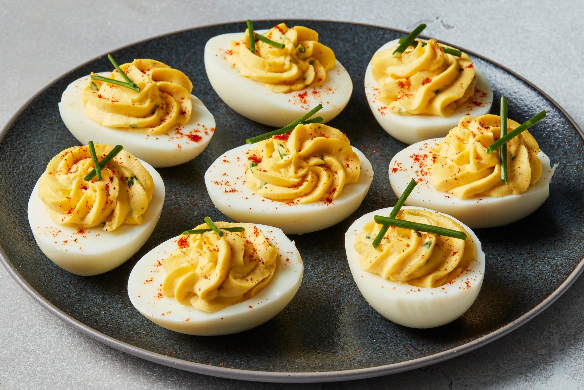 A dark platter of deviled eggs, with swirled fillings and topped with chives and paprika, sits on a light gray background.