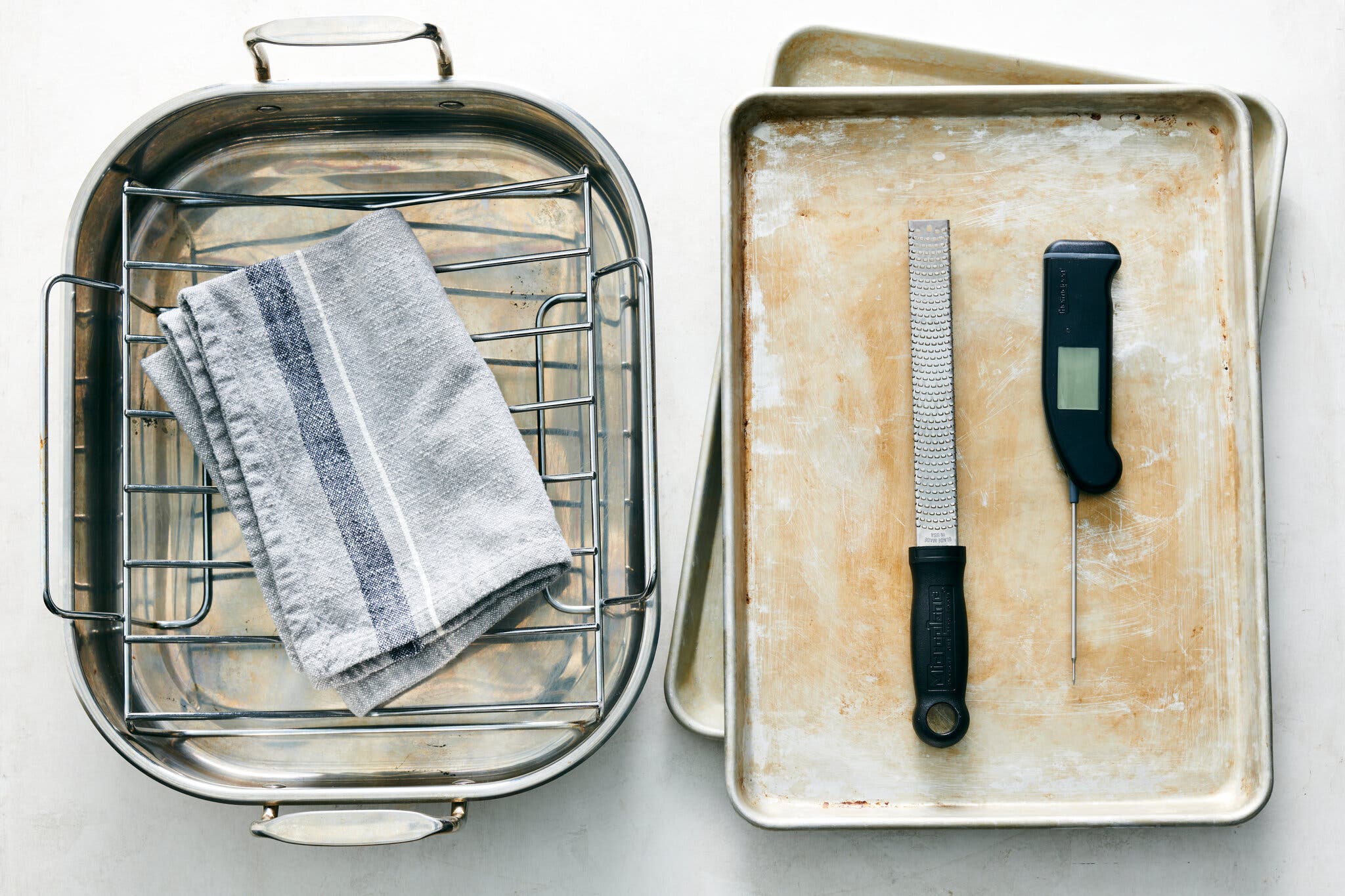 An overhead image of a roasting pan with a rack inside, a tea towel, two sheet pans, a grater and a thermometer.