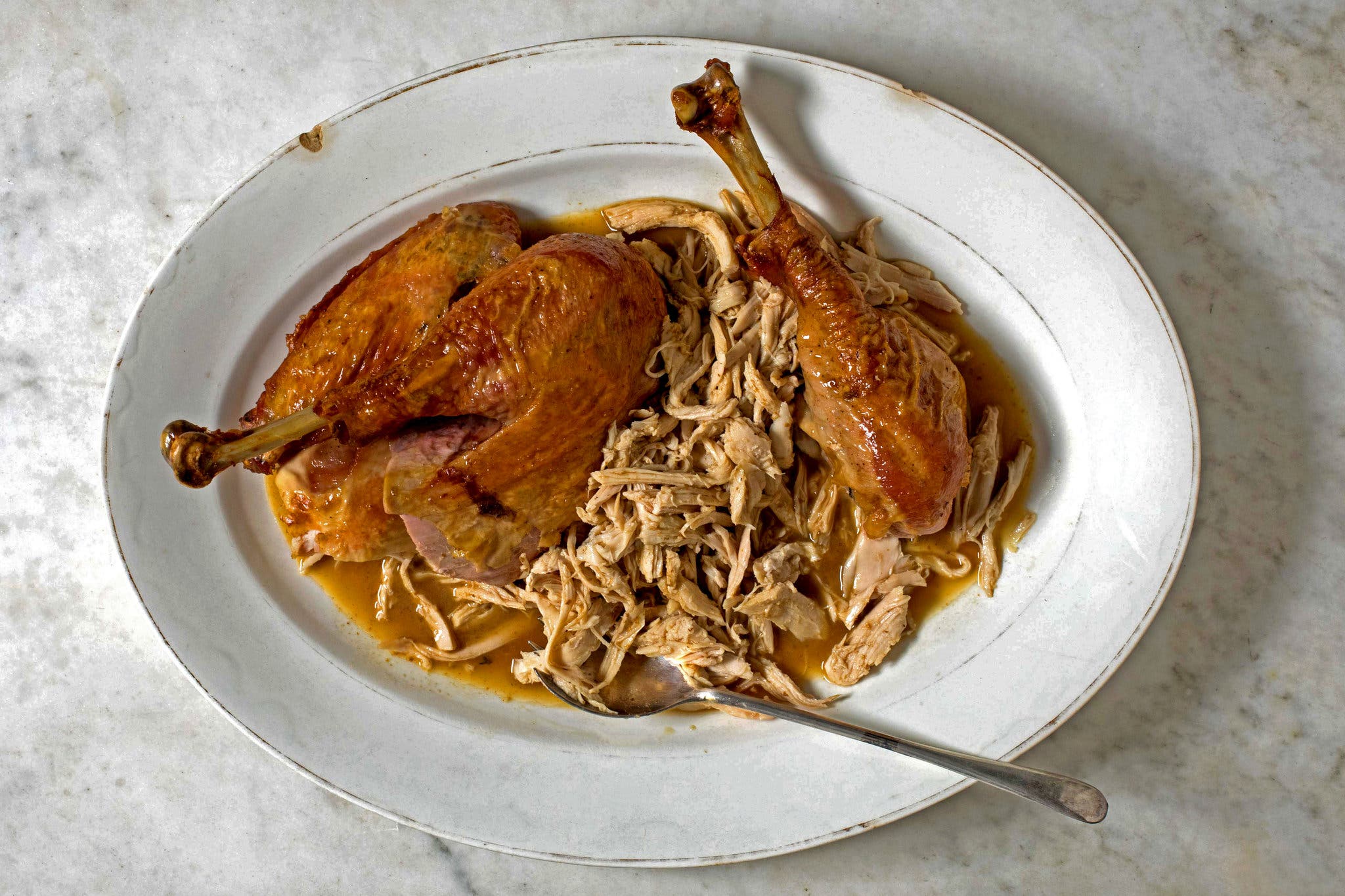 Two turkey legs and shredded turkey sitting in a sauce on a white platter, shot from overhead.