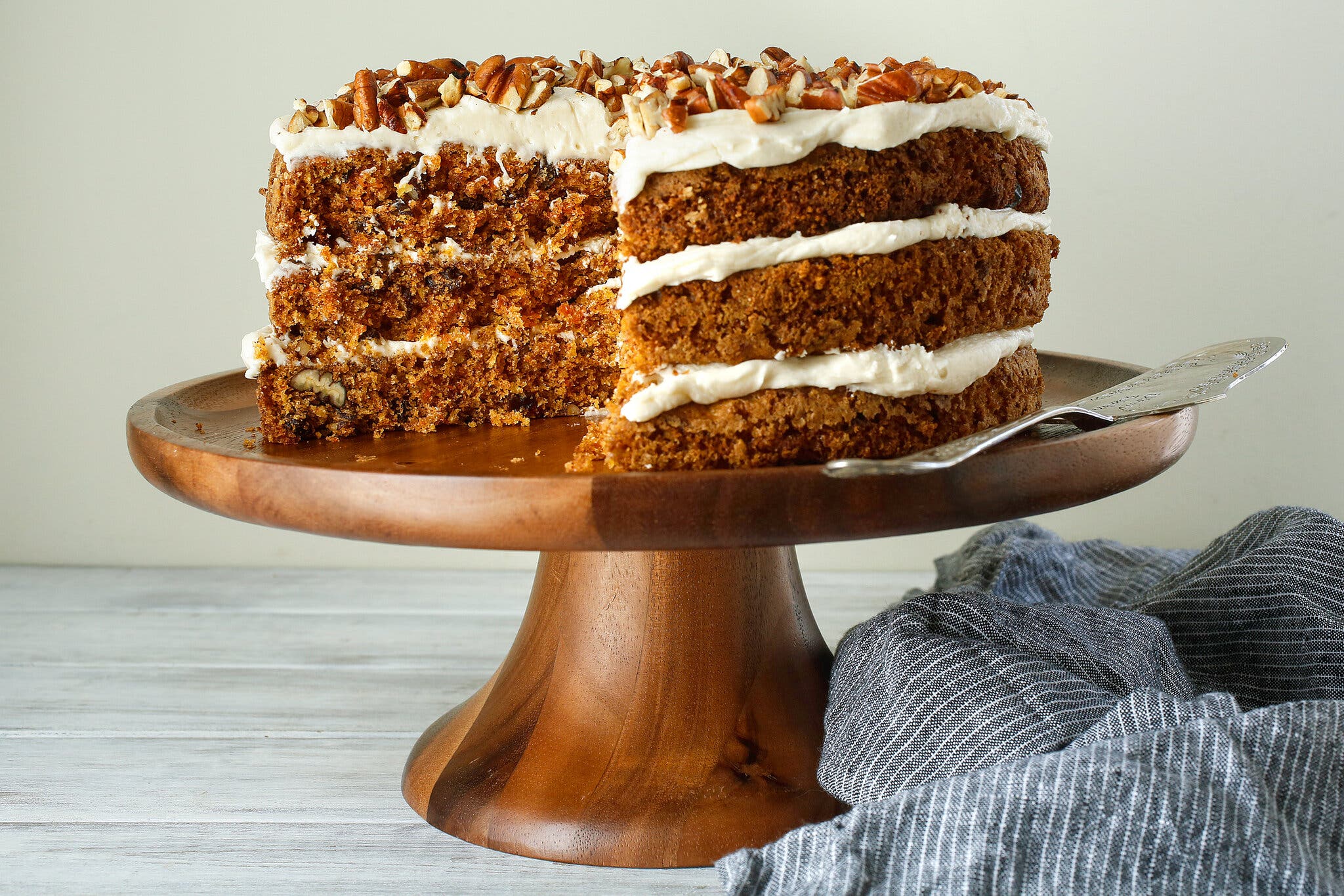 A layered carrot cake topped with nuts sits on top of a wooden cake stand. The edges are exposed to reveal the frosting between each layer. To the right is a blue striped fabric napkin.