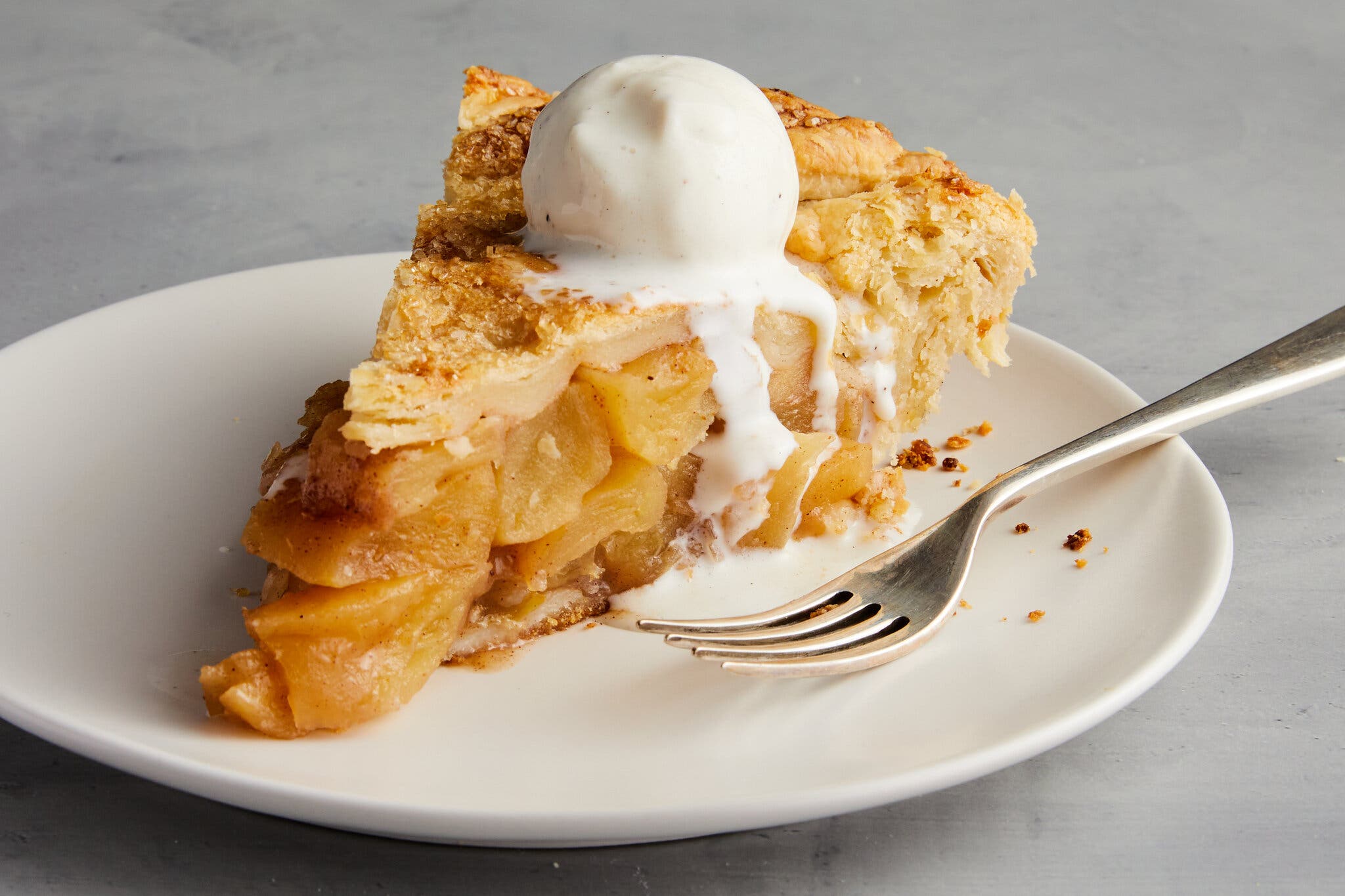 A piece of apple pie sitting on a white plate next to a metal fork is shot from the side to reveal its filling. A melting scoop of vanilla ice cream sits on top.