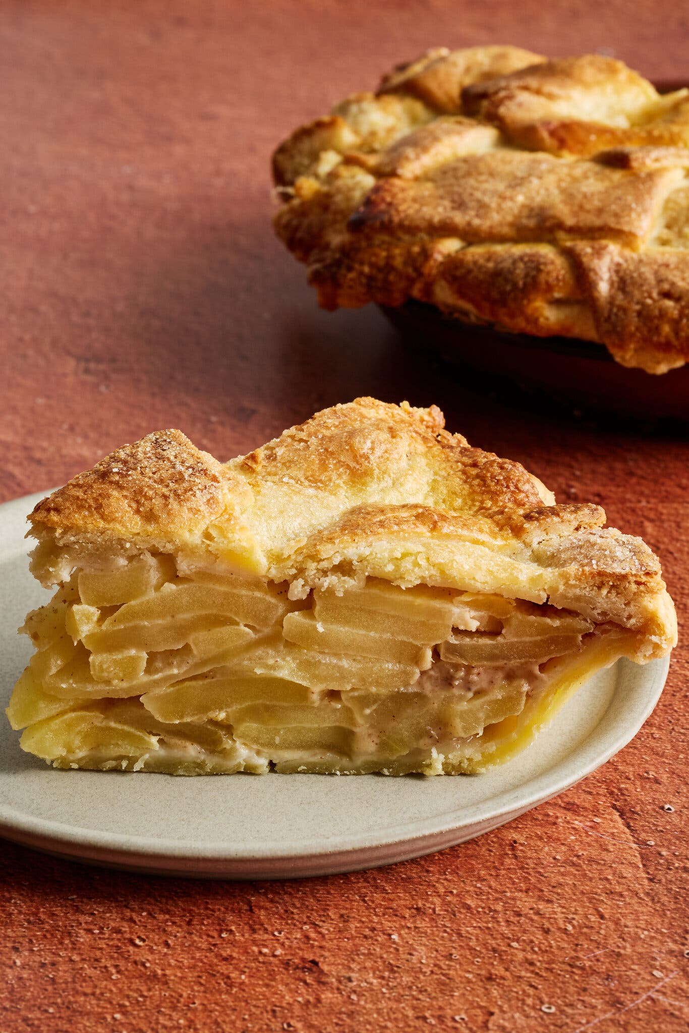 A slice of apple pie sits on a white plate, with the full pie half in frame behind it.