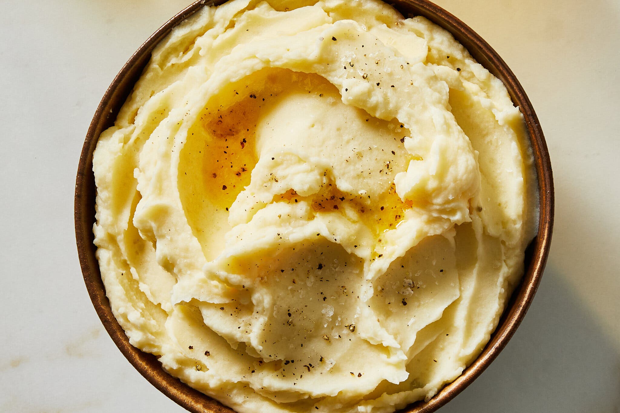 An overhead shot of a copper bowl of mashed potatoes, flecked with black pepper.