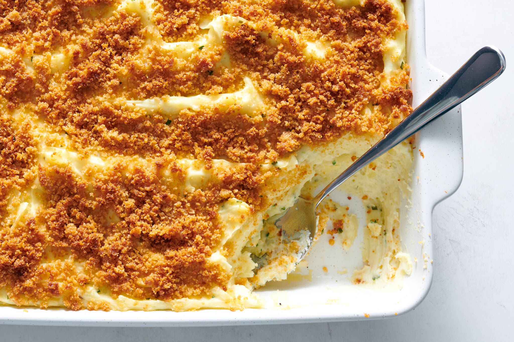 An overhead shot of a baking dish of mashed potato casserole, topped with bread crumbs. There is a spoon in the dish.