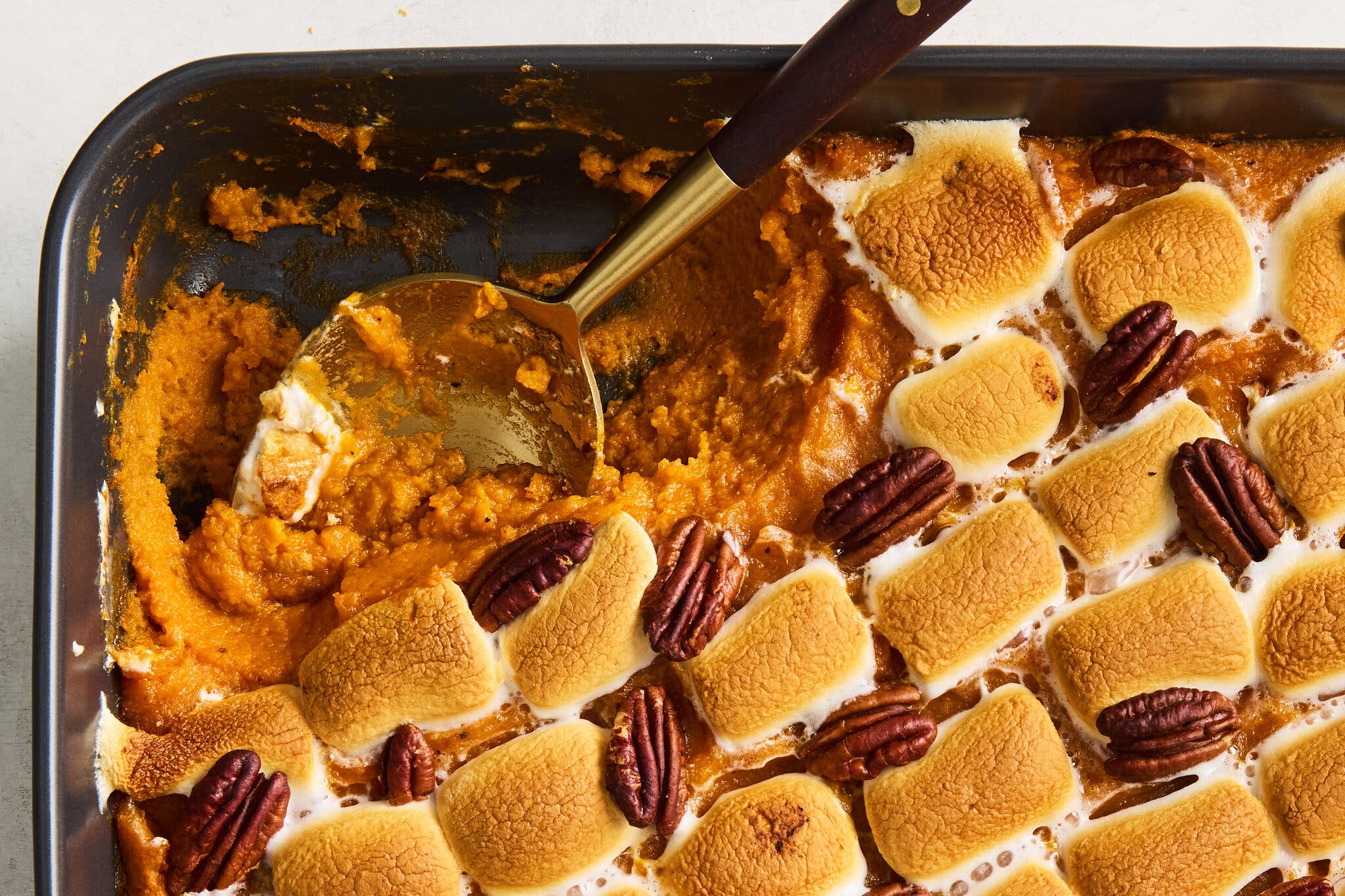 An overhead shot of a baking dish full of sweet potato casserole, topped with browned marshmallows and layered with pecans. There is a serving spoon in the dish.