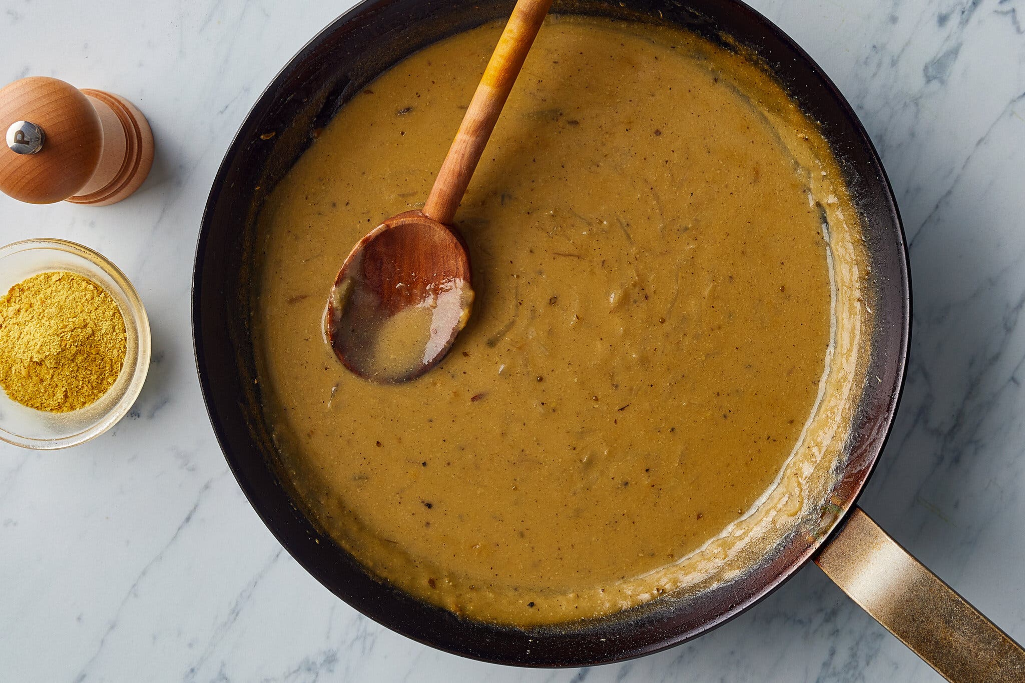 An overhead shot of a pan of gravy, with a wooden spoon in the pan. On the side are a small bowl of nutritional yeast and a pepper grinder.
