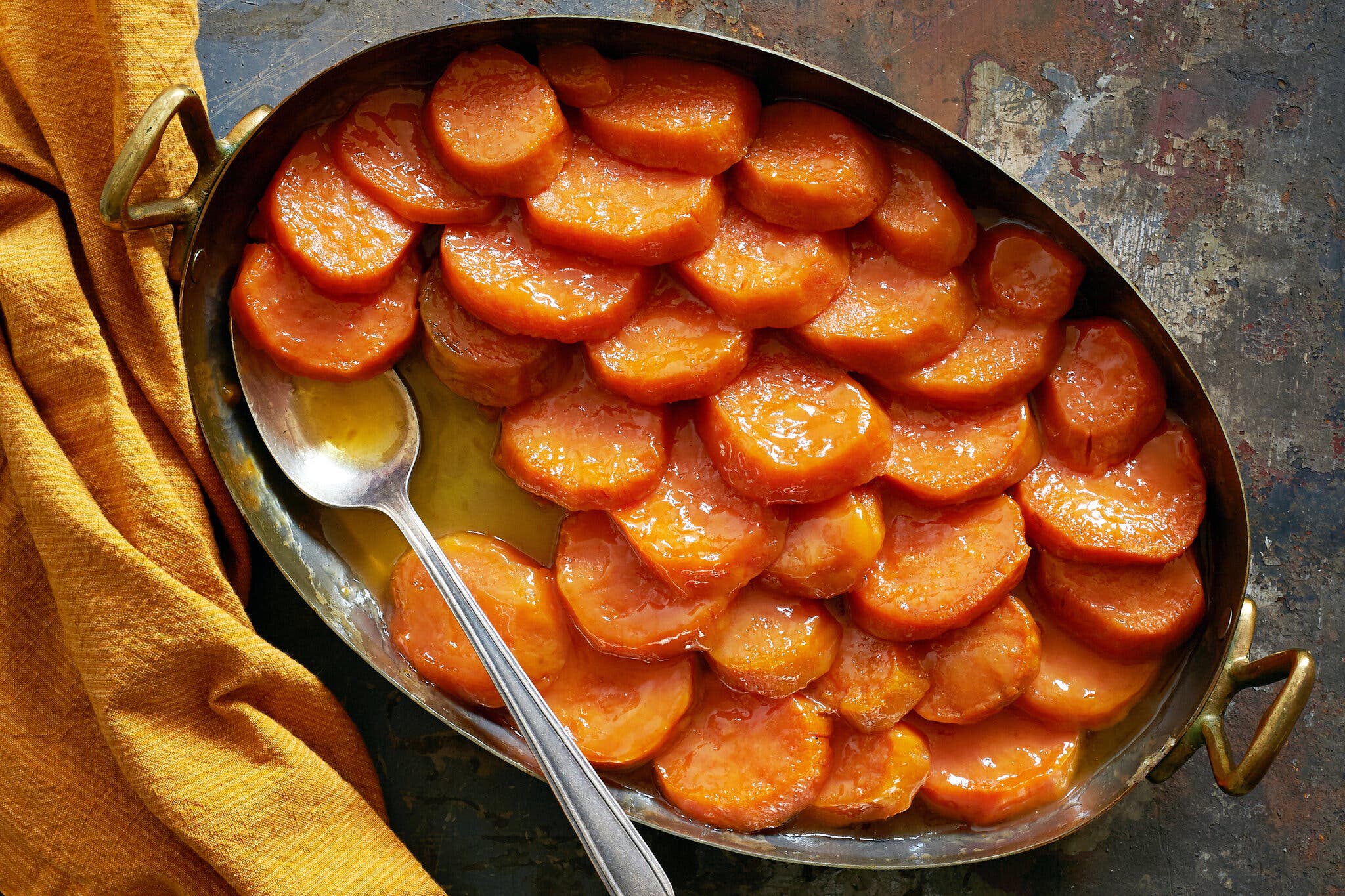 An overhead image of a brass-handled oval roasting dish filled with layers of glazed sweet potatoes. A large silver spoon is tucked into the potatoes. A rust-colored dish towel is bunched up in the left side of the image.