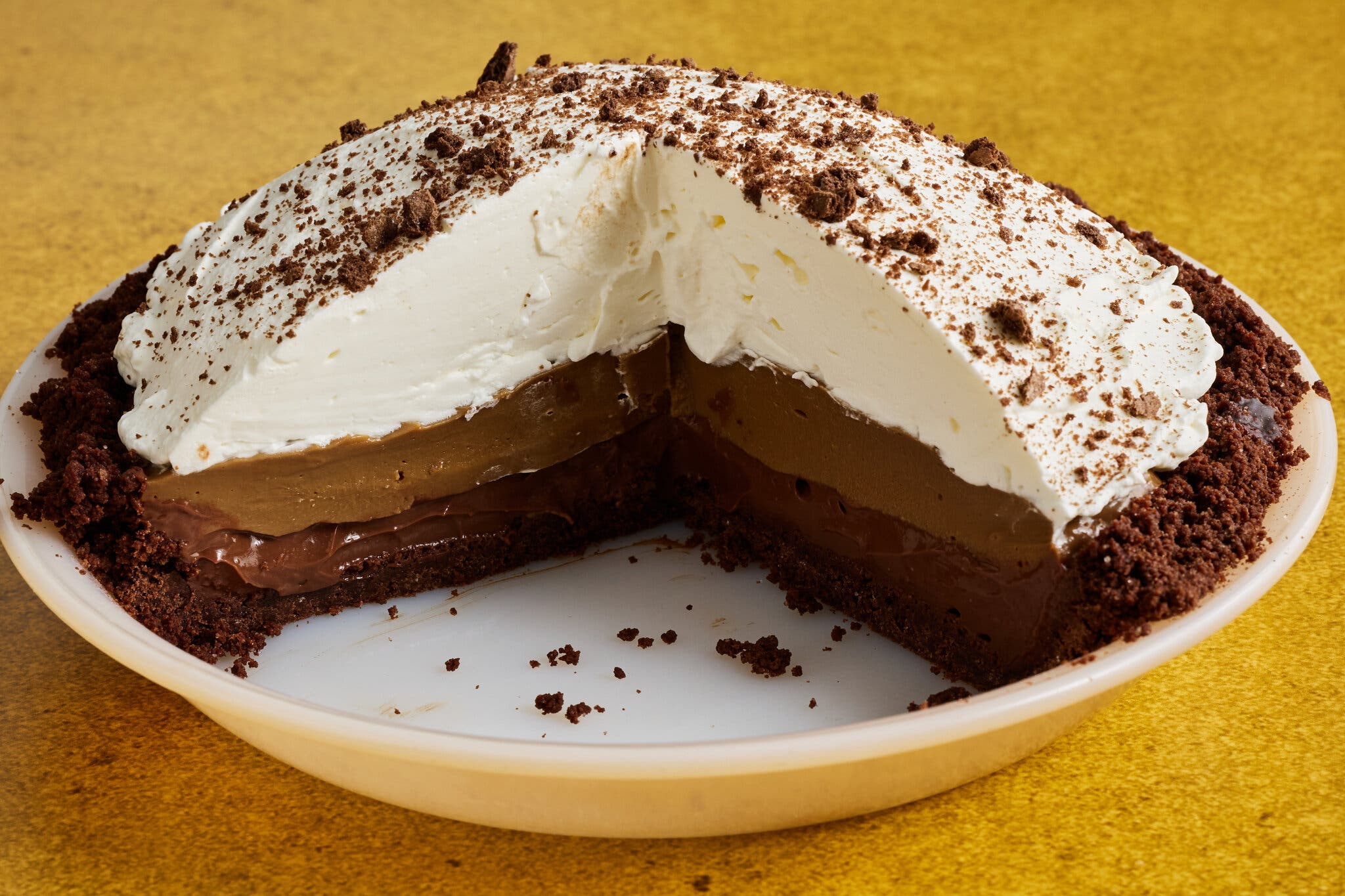A pie with a big slice missing, revealing dark brown crumbly crust, two brown filling layers and a thick topping of white whipped cream.