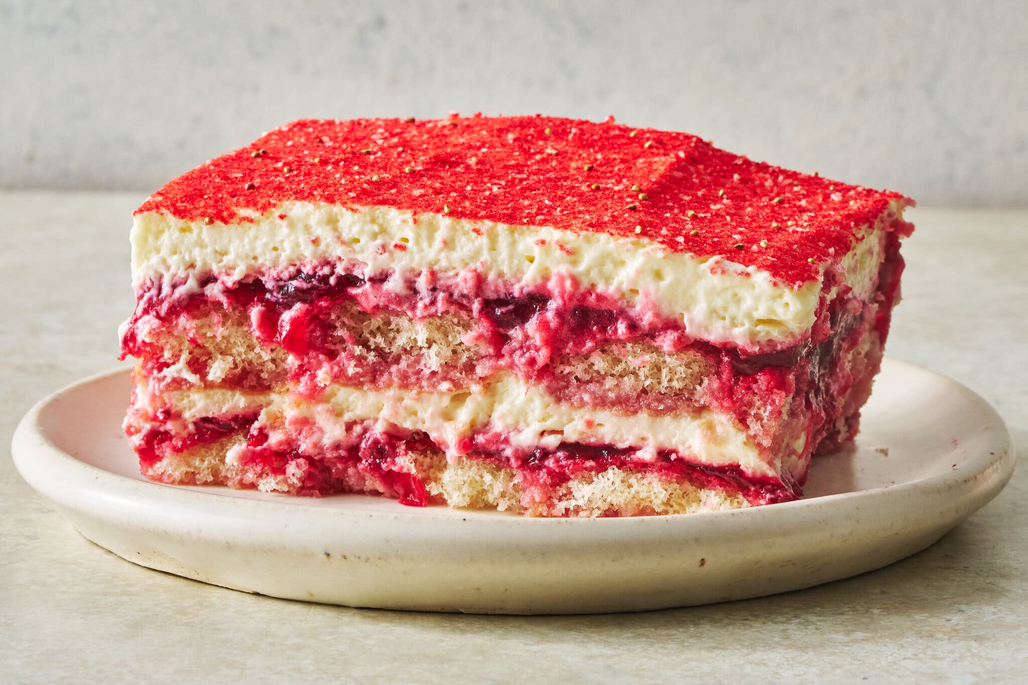 A slice of cranberry tiramisù with red fruit filling and white cream sits on a white plate. It is sprinkled in bright red powder.