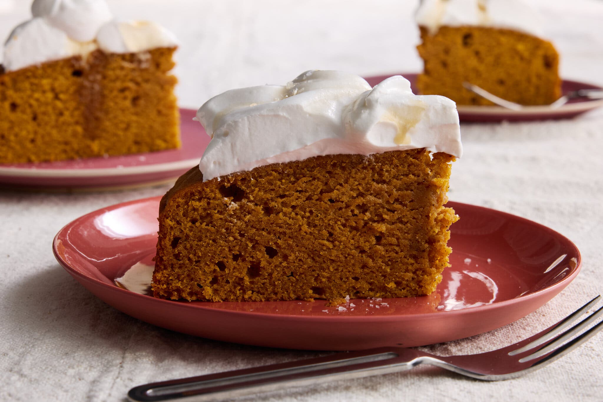 A slice of orange cake topped with white whipped cream sits on a red plate. Two other slices sit in the background.