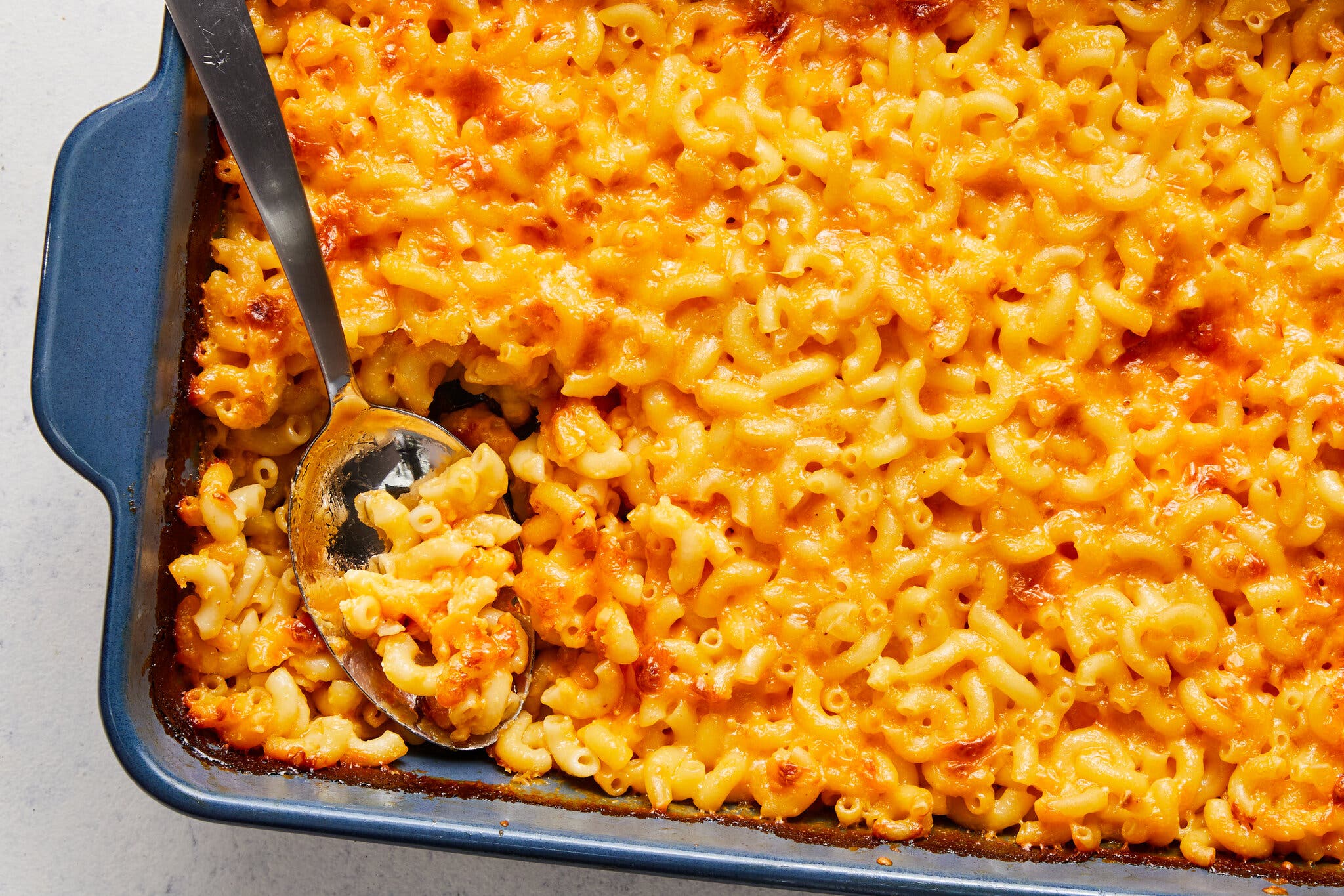 A close-up of baked macaroni and cheese with a bright orange, bubbly crust, served in a dark blue rectangular baking dish with a large silver spoon.