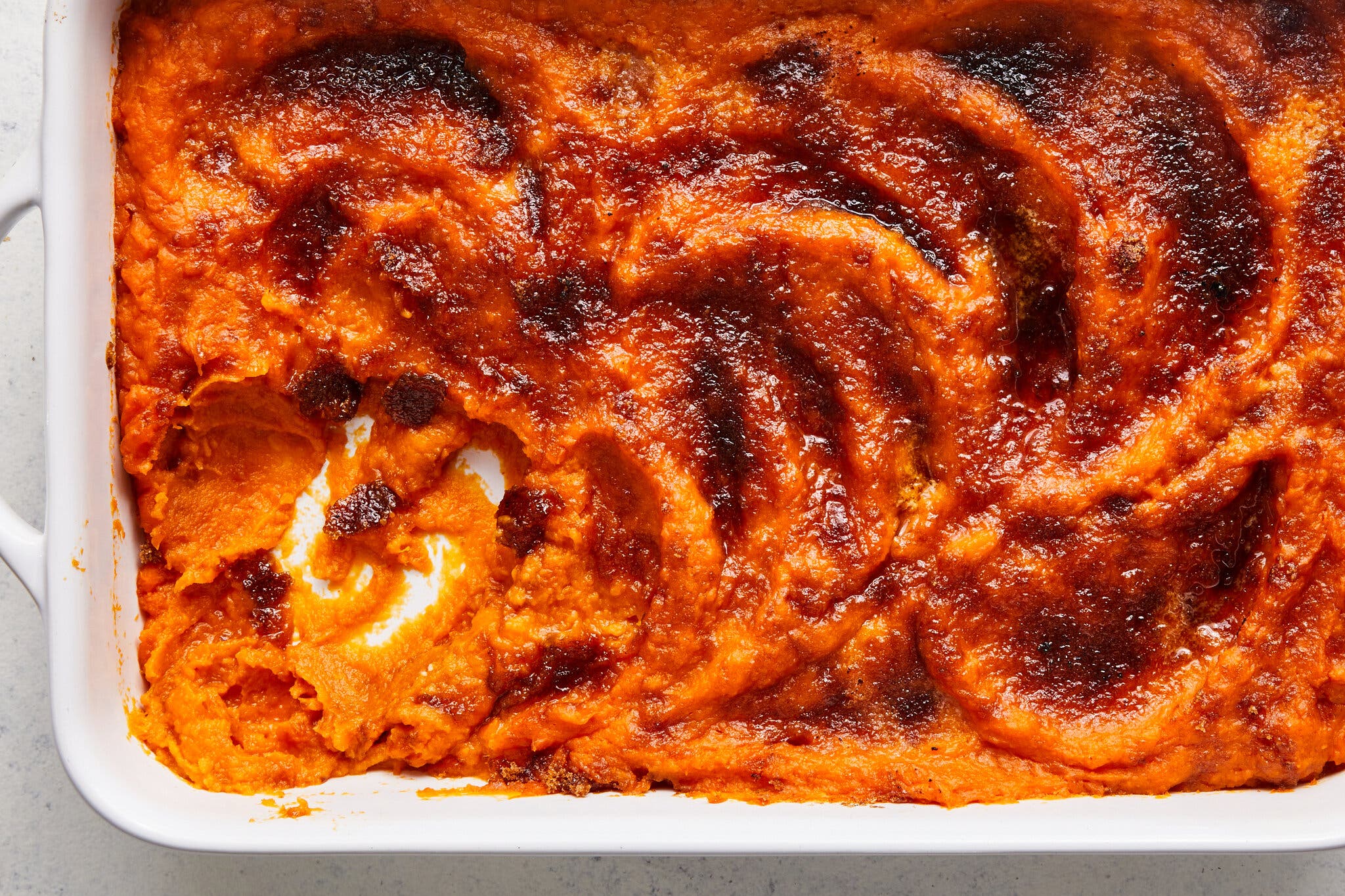 A close-up of a sweet potato casserole with a bright orange, mashed interior and a deeply caramelized, dark brown, sugary top baked in a white rectangular dish.
