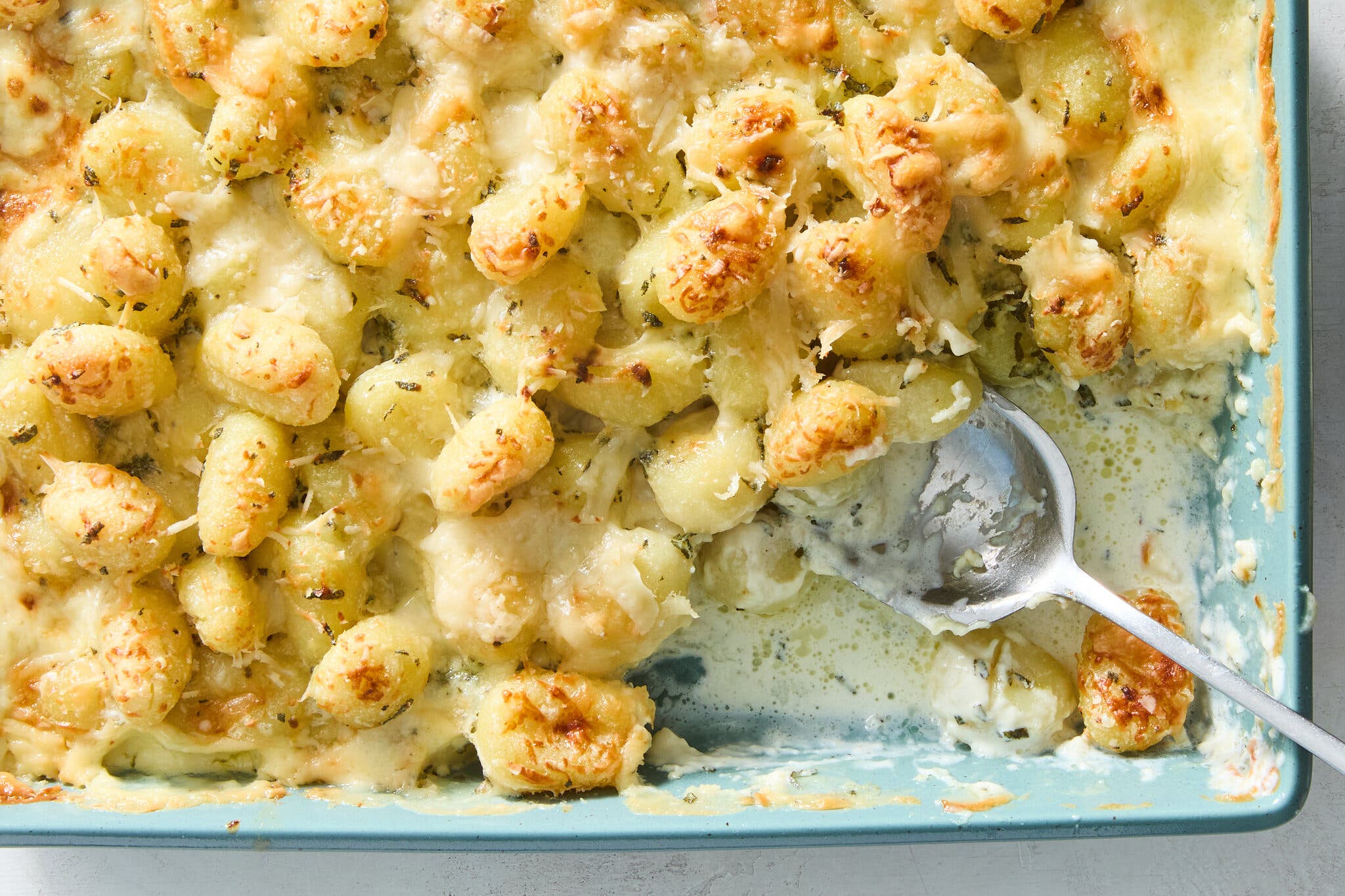 Baked potato gnocchi gratin with a creamy white sauce and golden-brown caramelized tops, served in a light blue baking dish with a silver spoon.