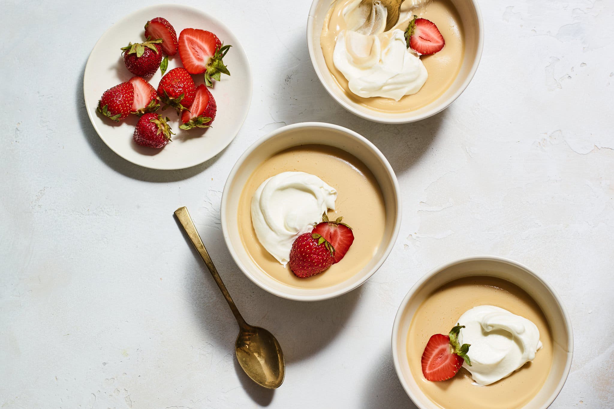 An overhead image of three bowls filled with butterscotch pudding, topped with dollops of whipped cream and strawberry halves. A gold spoon sits next to one of the bowls. A separate small plate is filled with strawberry halves.