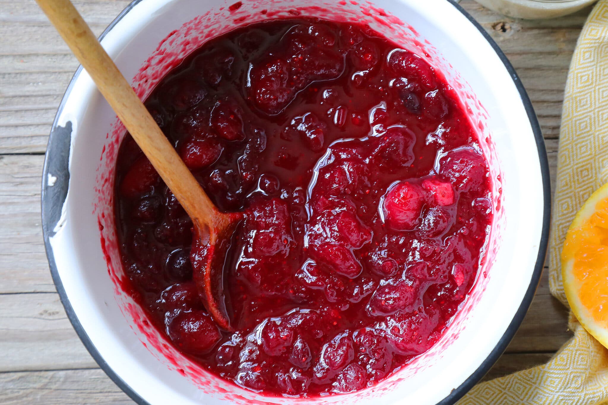 An enameled pot holds just-cooked cranberry sauce. To the right are halved, freshly squeezed oranges, and above is a bowl with sugar. 