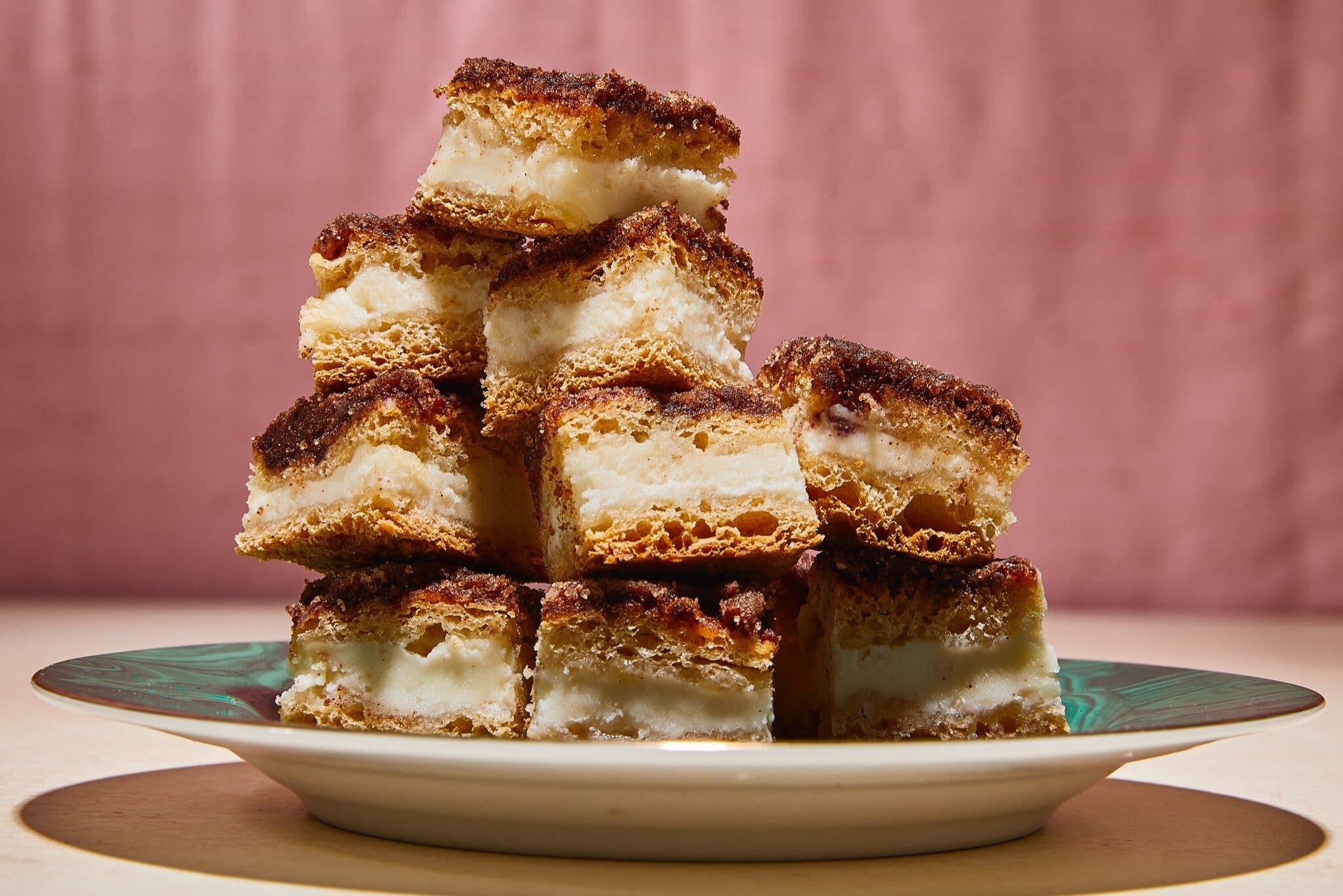 A plate of cheesecake bars stacked on top of one another. They have flaky dough layers as well as a creamy cheesecake layer in the center.