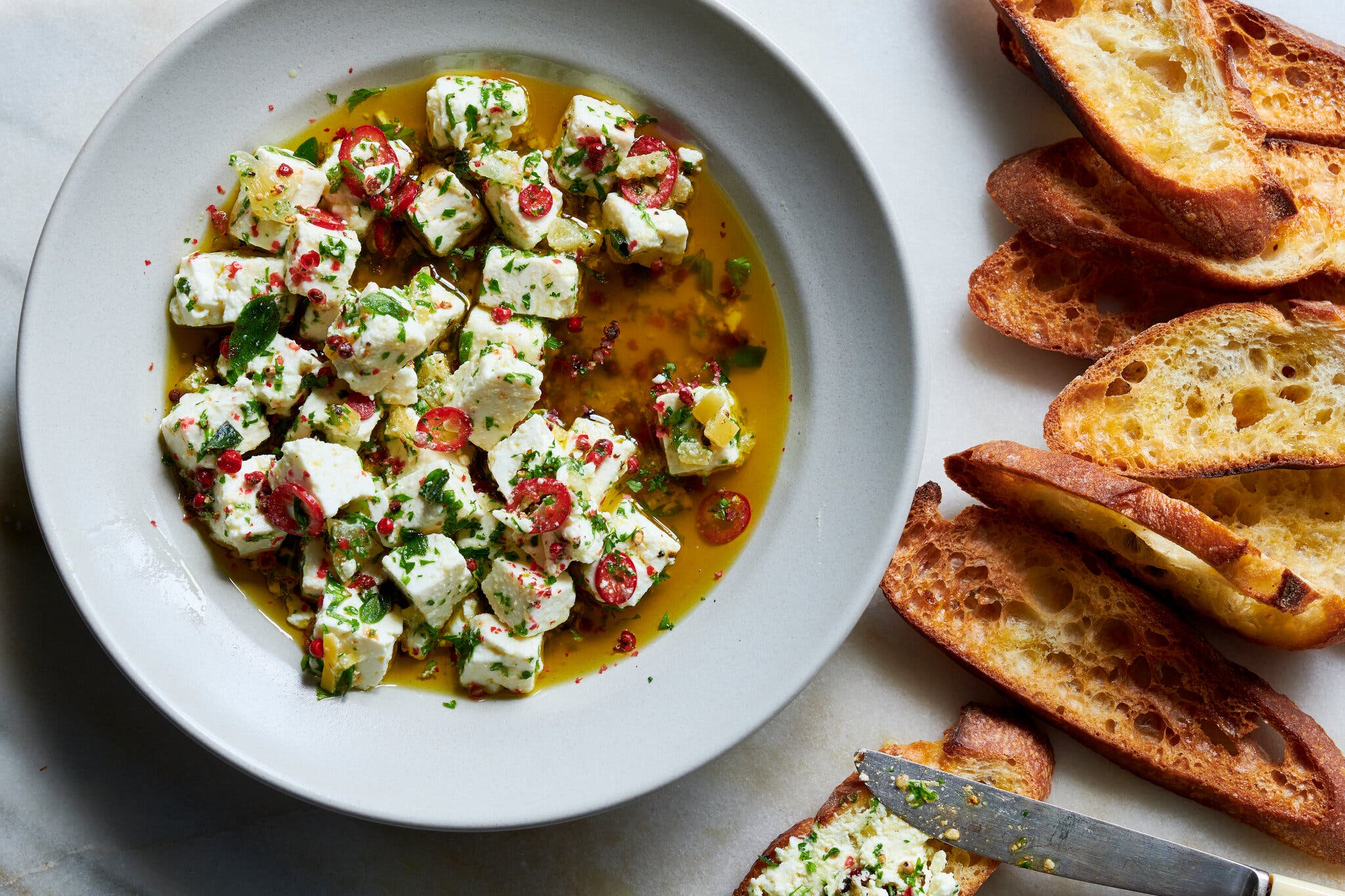 A white bowl filled with olive oil and feta cubes topped with herbs sits on a marble countertop. To the right are a number of baguette slices.
