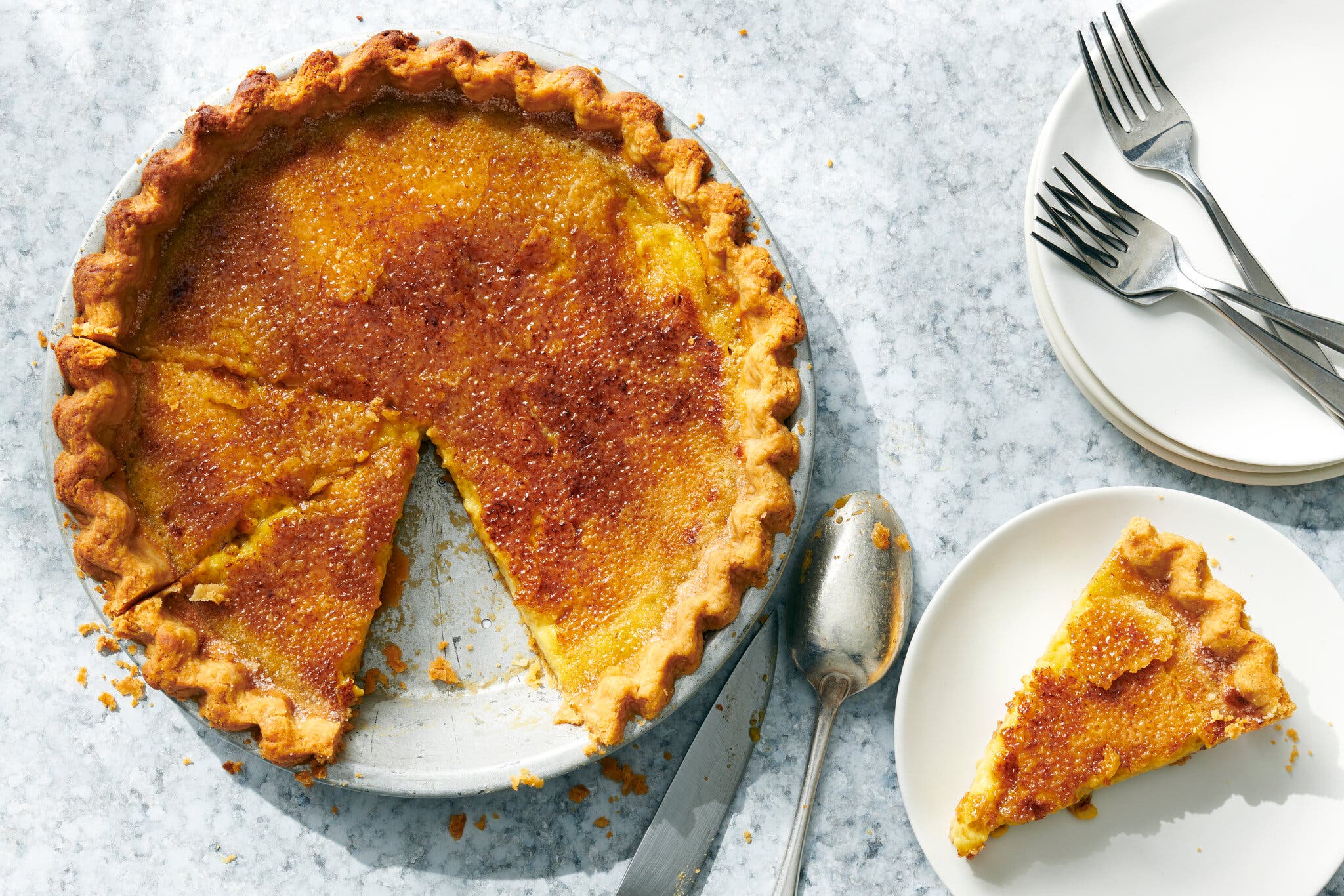 A burnished custard pie sits in a pie pan with a slice missing. To the right is a white plate holding a slice. Cutlery and plates are just off to the side.