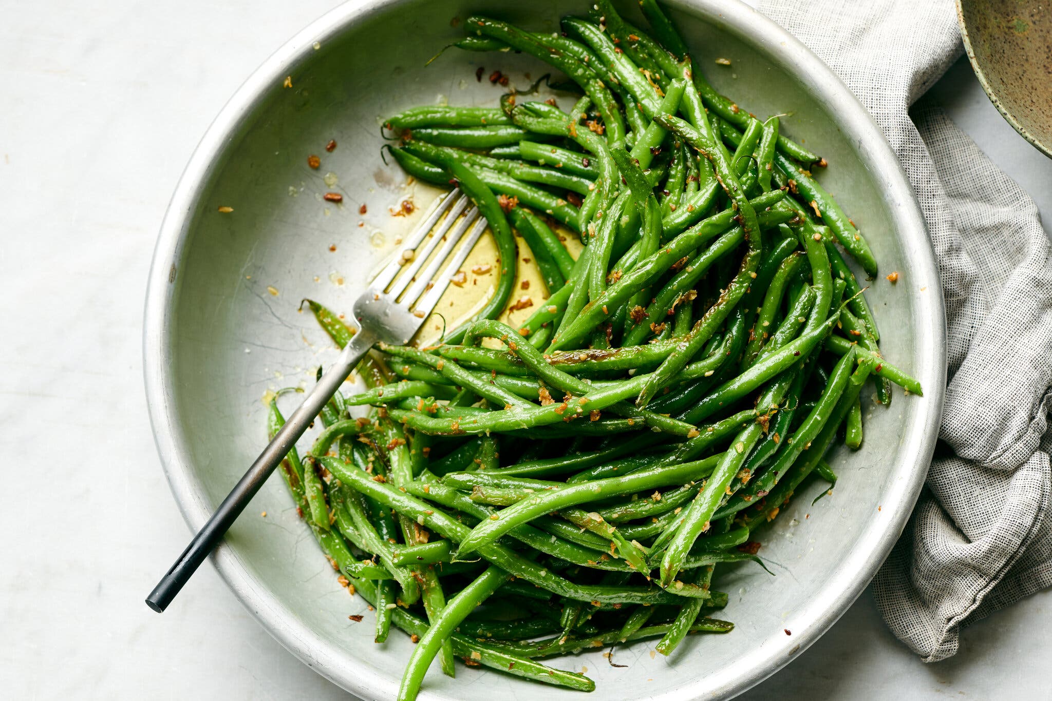 A bowl of green beans speckled with ginger and garlic is shot overhead. Just behind it is a textured gray napkin and a marbled background.