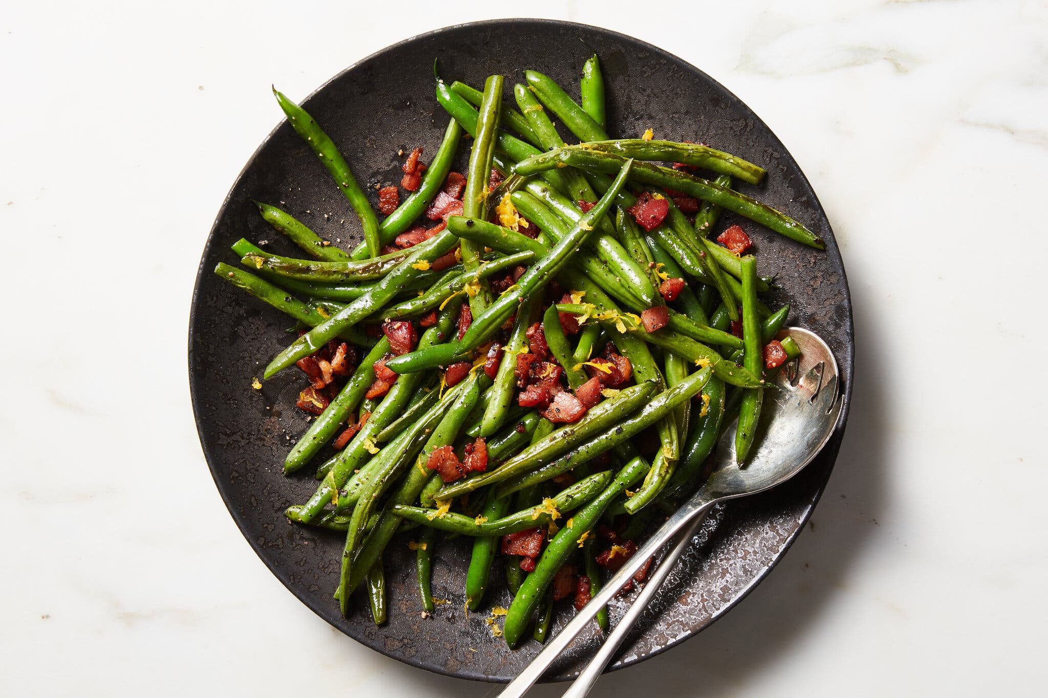 An overhead image of a black plate filled with green beans, chunks of pancetta and lemon zest. A silver serving spoon and fork are balanced on the right side of the plate.