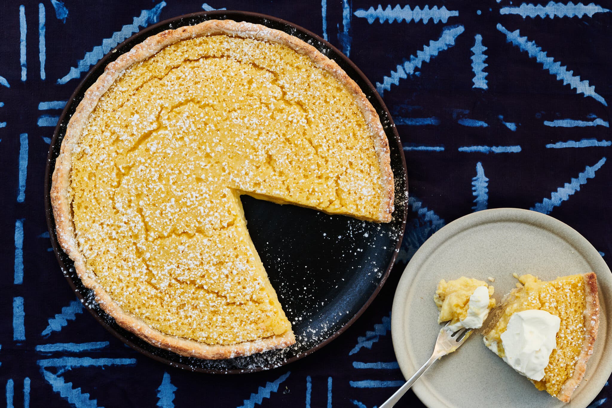 A yellow chess pie, dusted with confectioners’ sugar, in a dark pie dish sits against a light and dark blue patterned background. To the right is a plate holding a single slice topped with whipped cream.