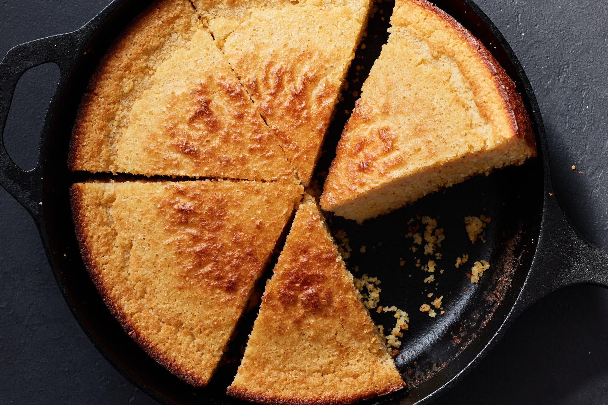 A cast-iron skillet filled with slices of cornbread sits against a dark gray surface.