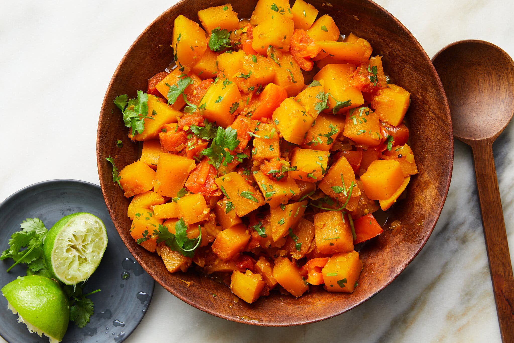 A wooden bowl holds cubed butternut squash topped with herbs. To the bottom left is a plate with spent lime halves and herbs.
