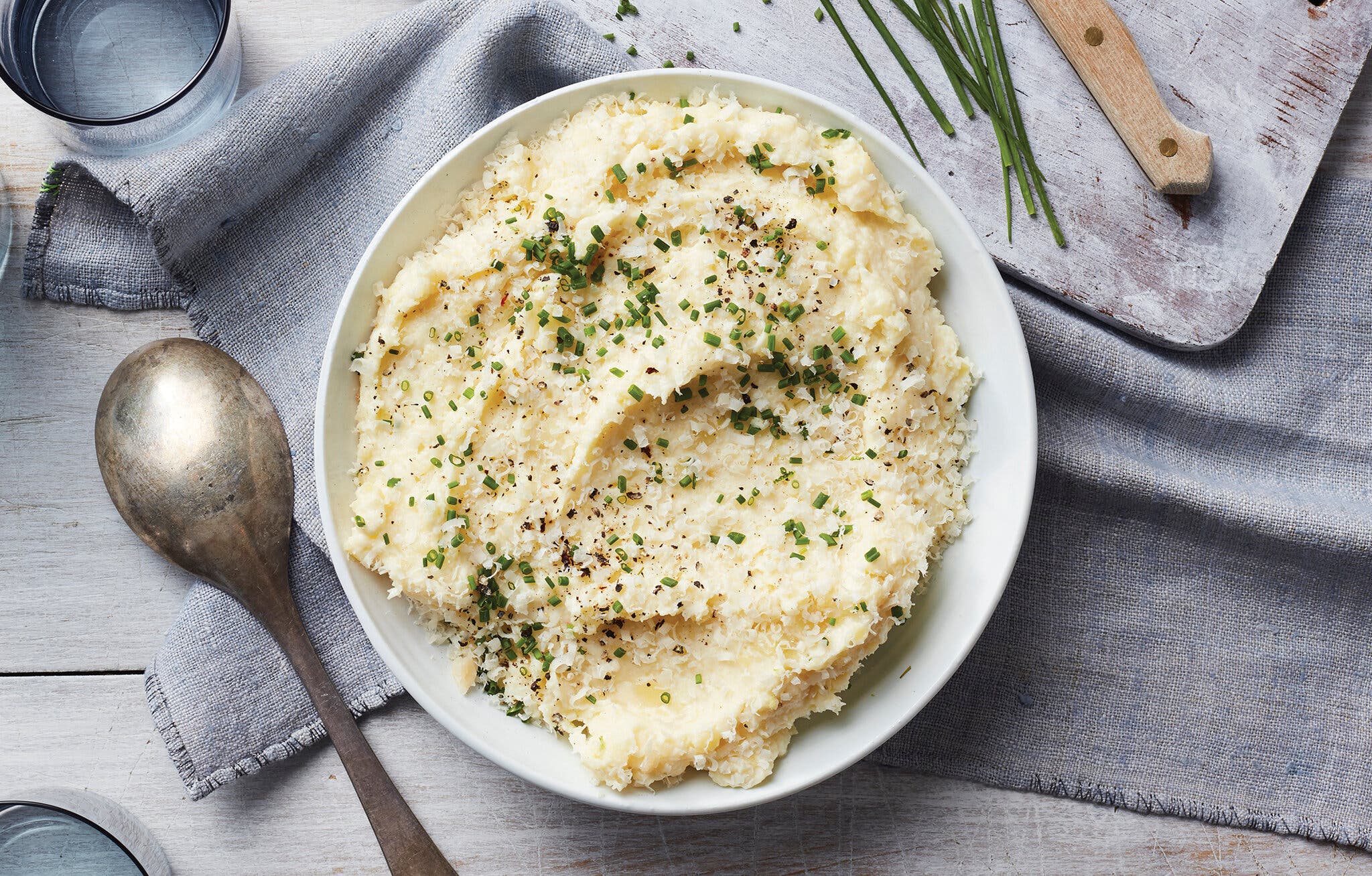 An overhead image of a white serving bowl on a gray tablecloth filled with mashed potatoes sprinkled with chopped chives and grated Parmesan cheese.