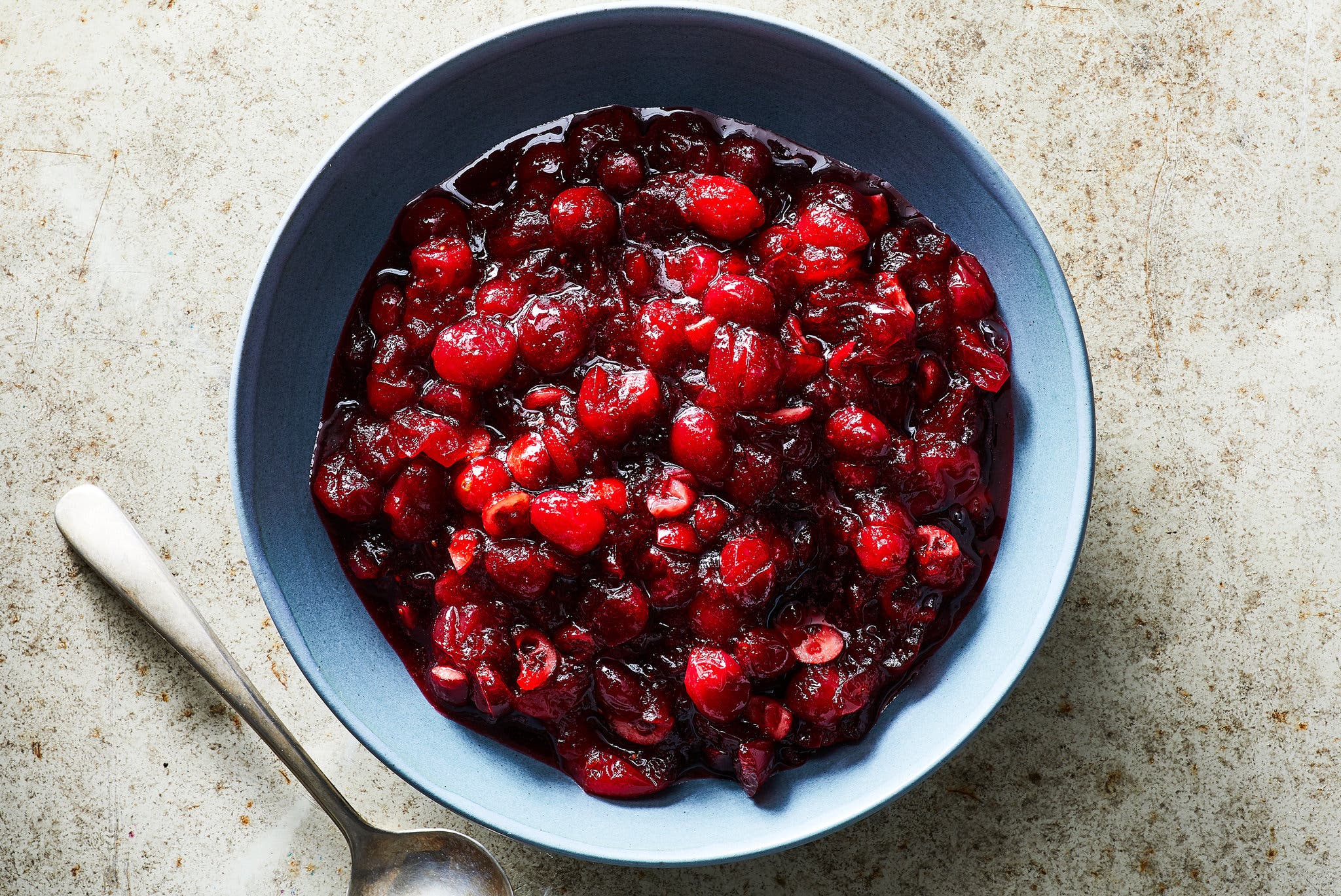 An overhead image of a slate blue bowl filled with cranberry sauce. A silver spoon sits to the left of the bowl.