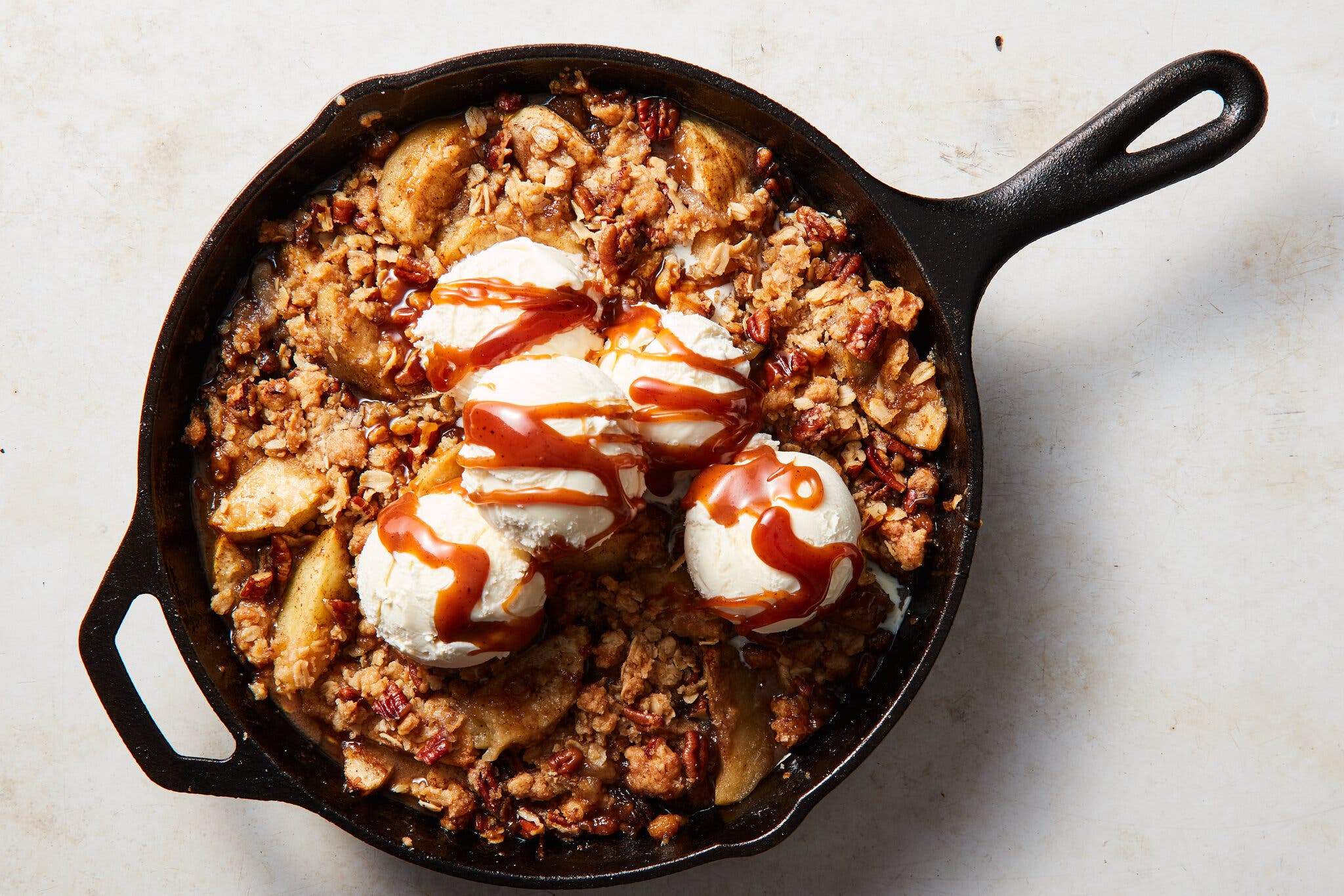 A cast-iron skillet filled with apples, a crumble topping and five scoops of vanilla ice cream drizzled with caramel sits on a light gray countertop.