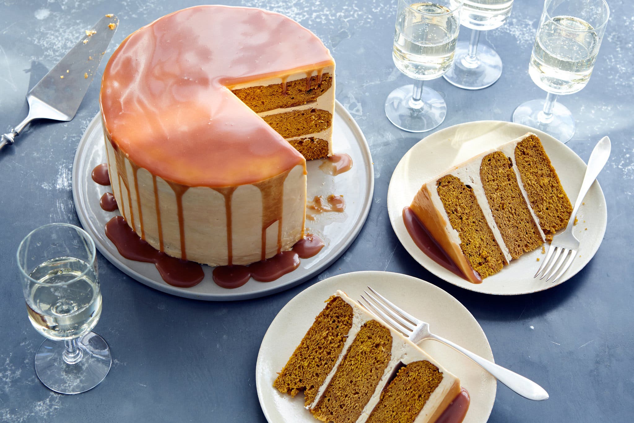 A tall, layered pumpkin cake covered with caramel sits next to two slices on white plates. Arranged around them are clear glasses filled with a light liquid.